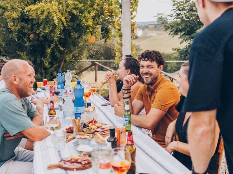 Lachende gasten aan een lange tafel buiten op agriturismo Vista Fermo, met planken vol Italiaanse vleeswaren, kaas, olijven en grissini, en het heuvelland op de achtergrond