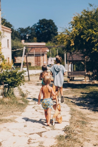 Drie kinderen in zwemkleding lopen over een stenen pad naar de schommel op agriturismo camping Vista Fermo, op een zonnige zomerdag