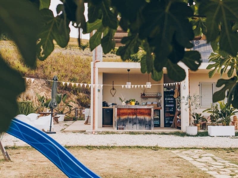 Blauwe glijbaan op het speelveld van agriturismo camping Vista Fermo, met daarachter de bar met vlaggetjes, gezien onder de bladeren van een vijgenboom