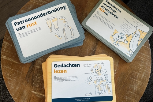 Drie stapels NLP Doe-Kaarten met geïllustreerde oefeningen over communicatie, gedachten en gedrag, uitgestald op een houten tafel.