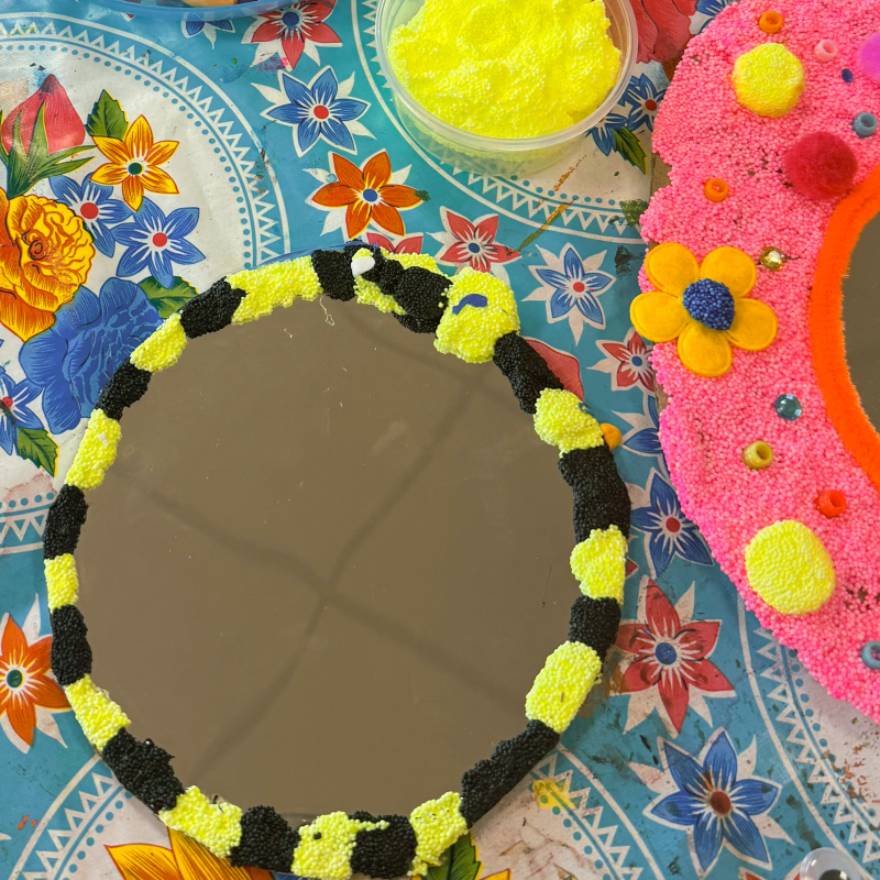 Workshop spiegeltjes versieren — overzicht resultaten Overzicht van versierde spiegeltjes op een kleurrijk tafelkleed — zwarte sterrenhemel, roze bloemen, geel-zwart gestreept en materialen