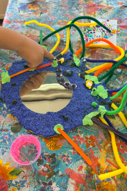 Blauw monster-spiegeltje met chenilledraad — in de maak Kind werkt aan een blauw spiegeltje met groene foamklei-accenten en kleurrijke chenilledraad die als tentakels omhoog steken
