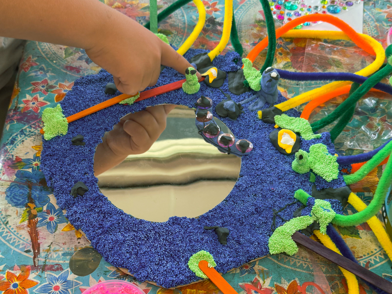 Kind werkt aan een blauw spiegeltje met groene foamklei-accenten en kleurrijke chenilledraad die als tentakels omhoog steken