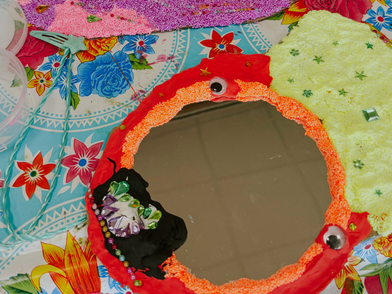 Twee versierde spiegeltjes naast elkaar — een geel-roze-paars exemplaar met strass en een rood-groen exemplaar met foamklei en wiebeloogjes