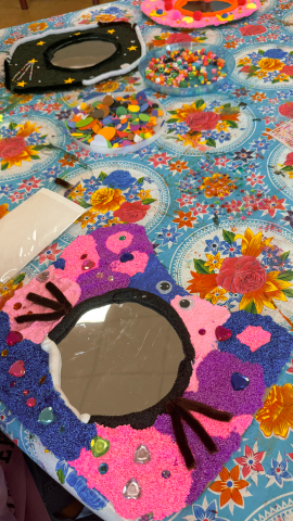 Werktafel met meerdere versierde spiegeltjes in verschillende stadia — foamklei in blauw, roze en paars, wiebeloogjes en chenilledraad