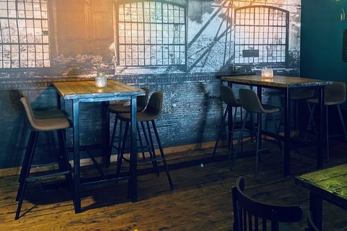 Hoge houten tafels met grijze barkrukken voor een muurschildering van oude fabrieksramen in Café Persee in Deventer