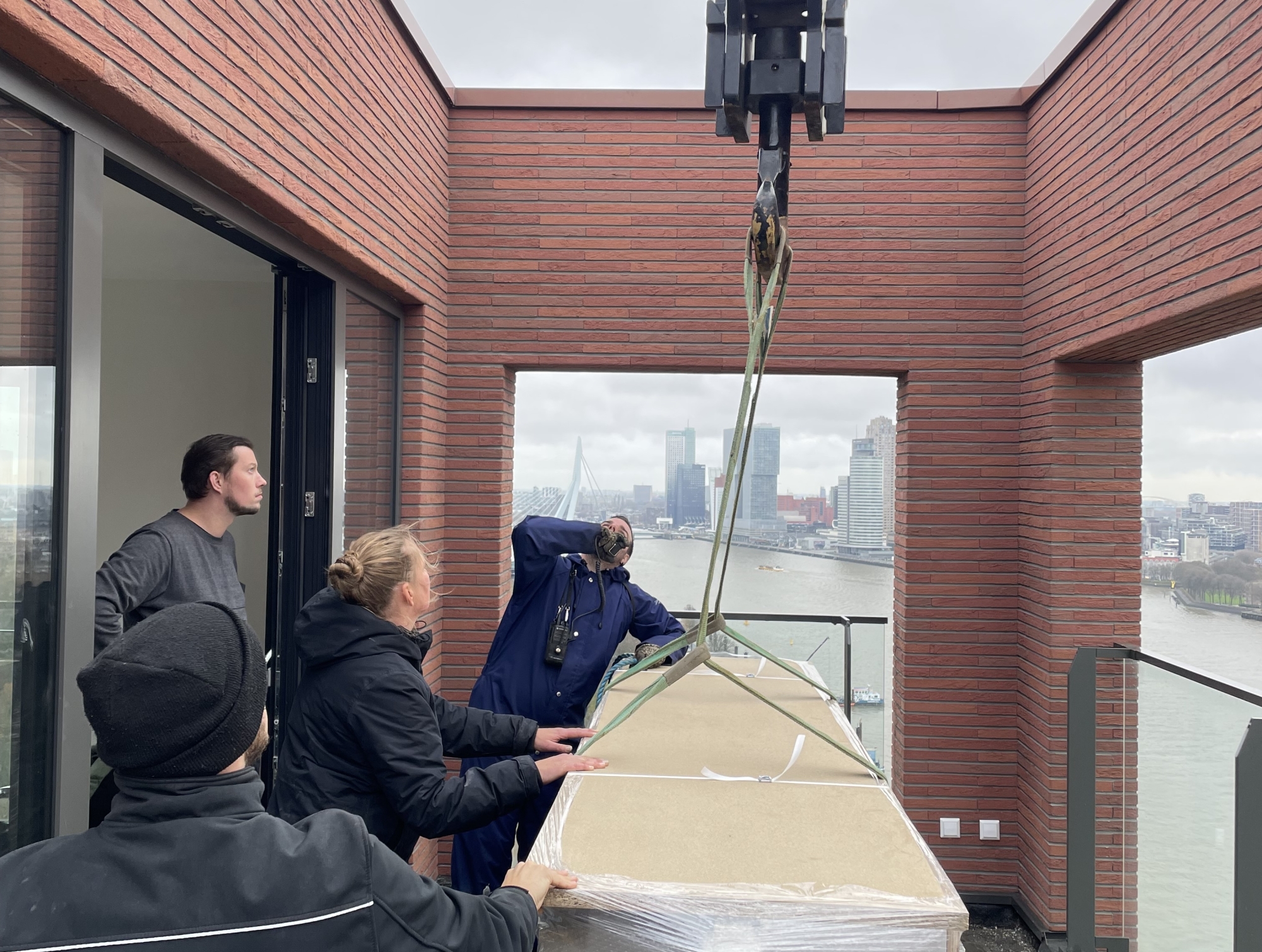 Maatwerk hijsen naar een penthouse in Rotterdam Maatwerkmeubel wordt met een kraan op het dakterras van een penthouse in Rotterdam gehesen, met zicht op de Maas