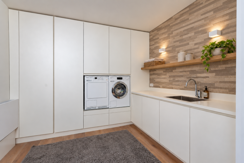 Wasruimte met witte kasten, een verhoogde wasmachine en droger, een spoelbak en wandverlichting boven een houten plank